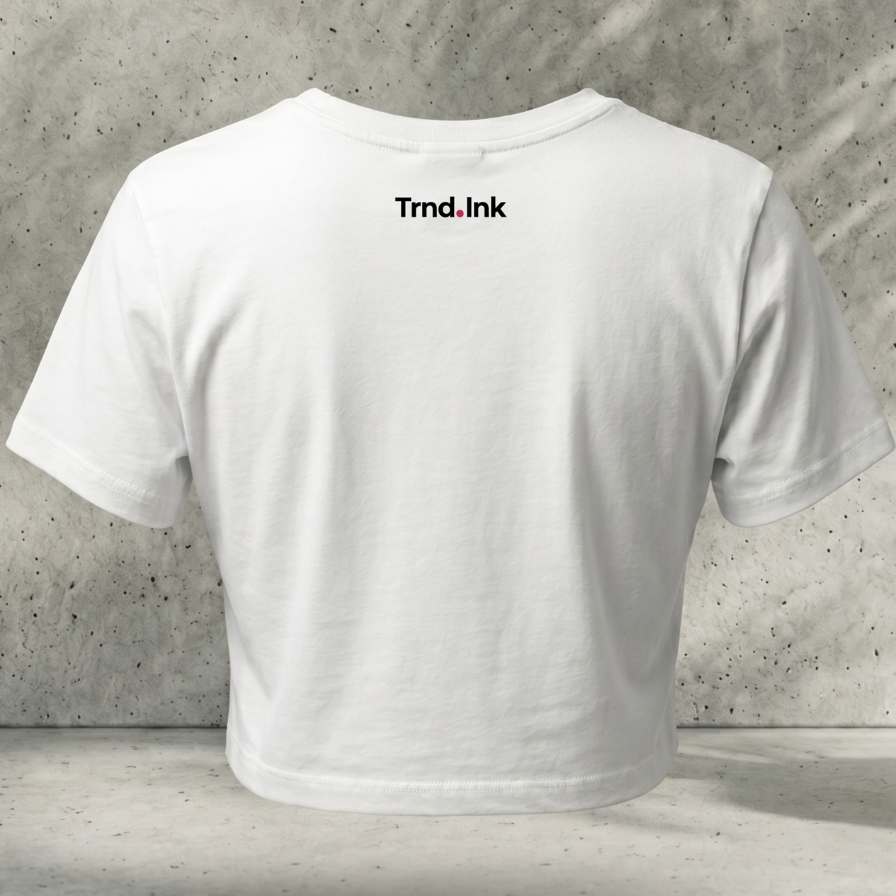 Vista trasera de un crop top blanco de algodón con el logotipo Trnd.Ink impreso en la parte superior central de la espalda, sobre un fondo de pared de concreto con textura.