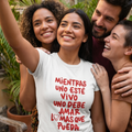 Mujer joven sonriendo y tomando una selfie al aire libre con un grupo de amigos, vistiendo una camiseta blanca con el mensaje estampado en letras rojas "Mientras uno esté vivo uno debe amar lo más que pueda