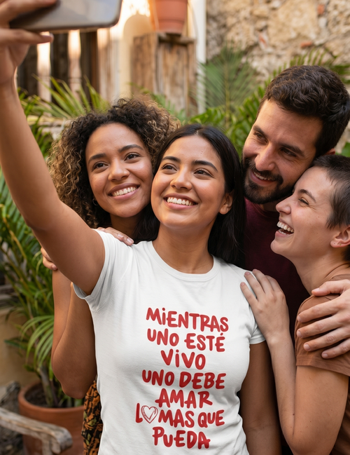 Mujer joven sonriendo y tomando una selfie al aire libre con un grupo de amigos, vistiendo una camiseta blanca con el mensaje estampado en letras rojas "Mientras uno esté vivo uno debe amar lo más que pueda