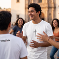 Un hombre joven de cabello rizado se ríe y conversa con un grupo de amigos en una plaza al aire libre frente a una iglesia histórica al atardecer. Lleva una camiseta blanca de la marca Trnd.ink con un estampado central minimalista de una cruz negra y la palabra "Fé" en caligrafía. Otra persona en primer plano muestra el logo de Trnd.ink en su espalda.