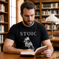 Hombre joven con barba leyendo un libro en una biblioteca clásica de madera, vistiendo una camiseta negra de manga corta con la palabra "STOIC" y el diseño de un busto de escultura romana fragmentado.