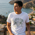Hombre joven posando al aire libre frente a un paisaje costero con montañas, vistiendo una camiseta blanca casual con el estampado vintage "La Révélation" y diseño de escudo heráldico.